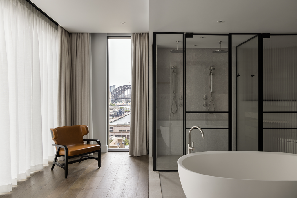 Image of Liberty Suite Bathroom with soaking tub and rain shower in the foreground, view of Sydney Harbour Bridge through the window in the background.