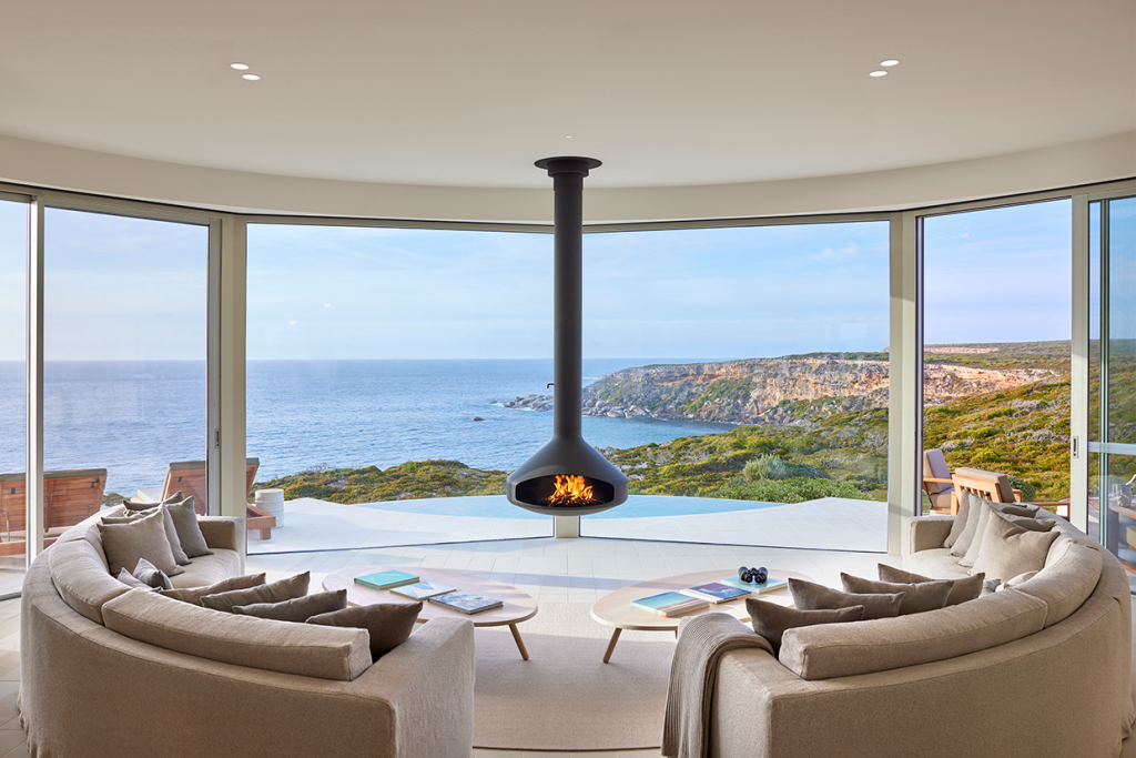 Ocean Pavilion Lounge with curved couches and freestanding fireplace in foreground, sweeping view of cliffs and ocean through the windows in the background.