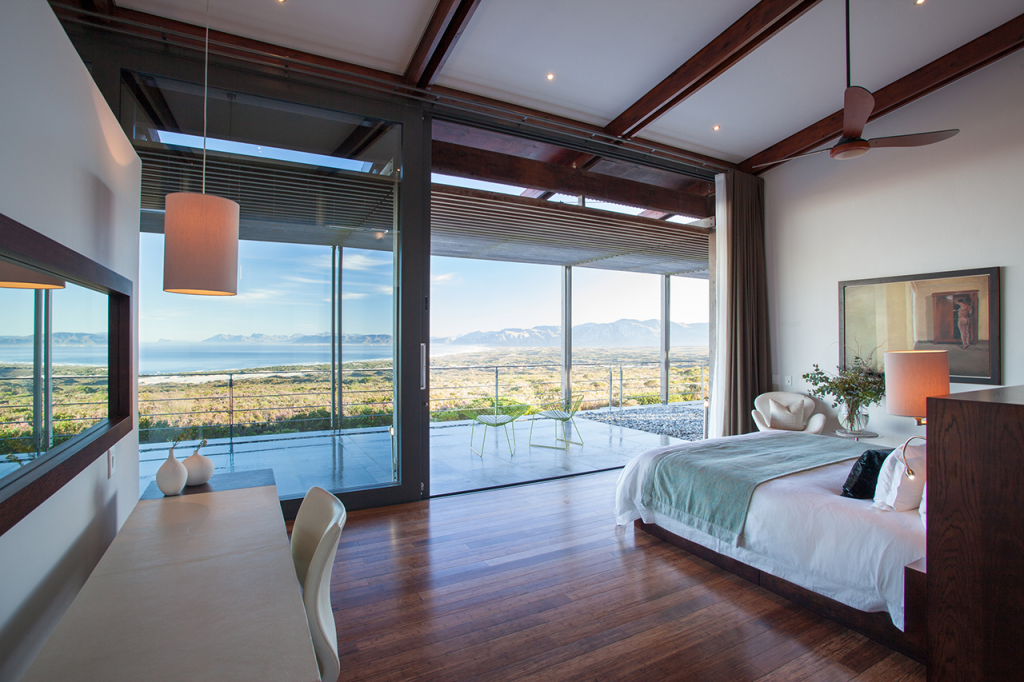 Image of a villa bedroom in the foreground with view of the balcony and the ocean and mountains beyond in the background.