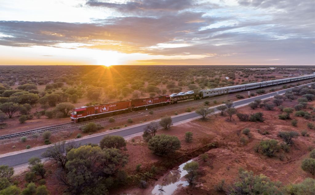 Image of The Ghan train rolling through the countryside with the sun setting in the background.